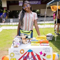 Person with glasses smiling with a table of prizes.